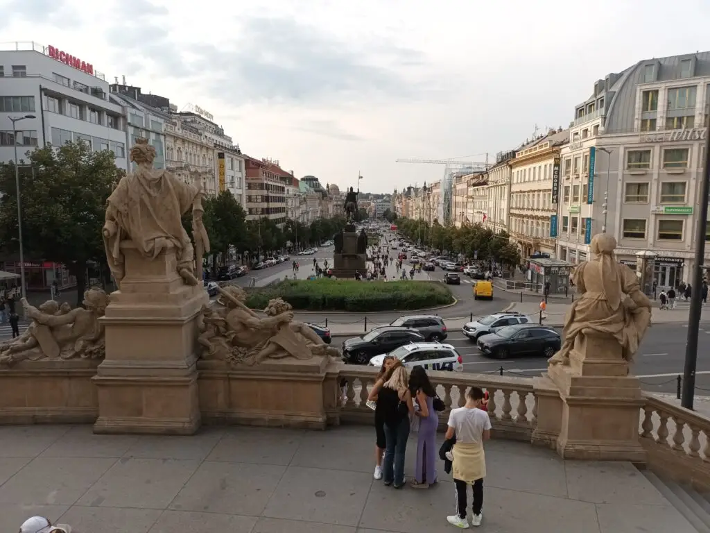 [:en]Wide street in Prague. In the foreground, people are looking at the statues.[:]