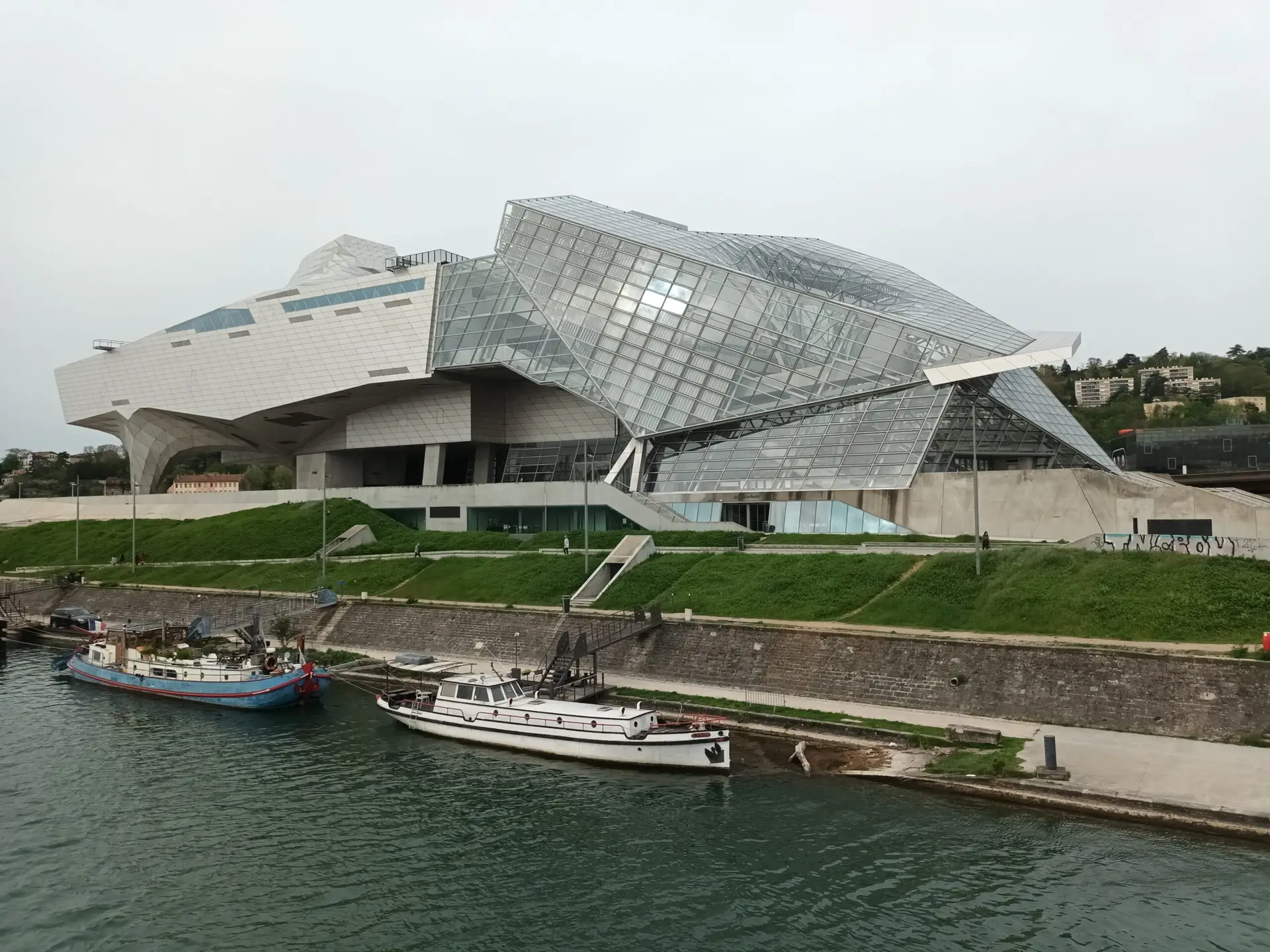 [:en]Museum of Confluences in Lyon. Curved roof structures. In the foreground, a canal with large boats along the shore.[:]
