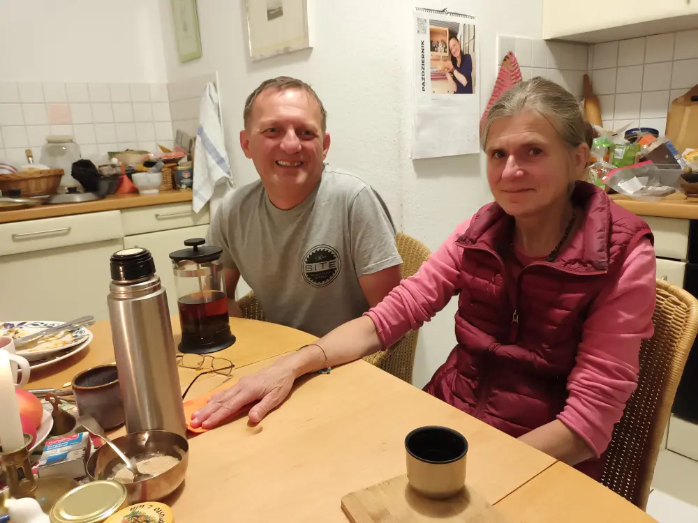 A man and a woman are sitting at the kitchen table and looking at the camera.